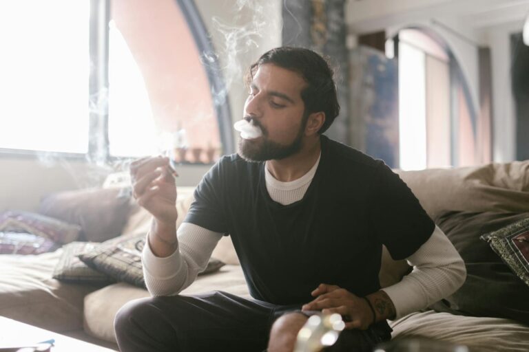A man with facial hair smokes indoors on a sofa, wearing a black shirt and white undershirt.