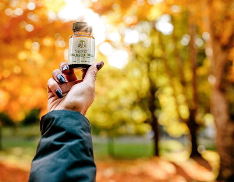 Hand with manicured nails holding CBD oil bottle in fall forest setting.